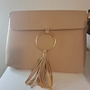 Beige axelväska med fransdetalj - Snygg beige axelväska i syntetmaterial med en stor ringdetalj och fransar på framsidan. Väskan har en dragkedja inuti för extra säkerhet. Perfekt för både vardag och festliga tillfällen! Kjedian kommer Inte med!