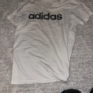 Vit t-shirt från Adidas - Säljer en vit t-shirt från Adidas i bomull. Den har en stor svart logga på framsidan. Perfekt för träning eller vardagsbruk. T-shirten är kortärmad och har en normal passform. Lite skrynklig men i bra skick! Brukar ha den som sovtröja