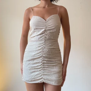 Champagnefärgad kortklänning med smala axelband - Säljer en elegant champagnefärgad kortklänning med smala axelband. Klänningen har en åtsittande passform och rynkade detaljer som ger en snygg effekt. Perfekt för sommarens fester eller en utekväll. Materialet är lätt och bekvämt.