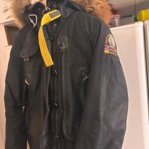 Svart parkajacka från Parajumpers - Säljer en svart parkajacka från Parajumpers med pälsfodrad huva och gul detalj vid halsen. Jackan har flera fickor och en robust dragkedja. Perfekt för kalla dagar.