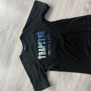 Svart t-shirt från Trapstar - Snygg svart t-shirt från Trapstar med ett coolt tryck i blå och vit nyans på framsidan. Perfekt för en avslappnad stil. T-shirten har en klassisk rund hals och korta ärmar. Priset kan diskuteras fraktar inom 12 timmar 📦🚚✅