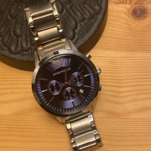 Emporio Armani herrklocka i silver - Snygg herrklocka från Emporio Armani med en stilren design. Klockan har en blå urtavla med flera sub-dials och datumvisning. Armbandet är i silverfärgat  vilket ger en elegant look. Perfekt för den som vill ha en tidlös accessoar. 