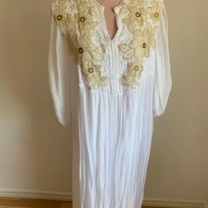 Vit långklänning med blommig spets - Elegant vit långklänning med vackra blommiga spetsdetaljer i guld på bröstet. Klänningen har en v-ringad design och långa ärmar, vilket ger en stilren och sofistikerad look. Perfekt för speciella tillfällen.