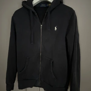 Mörkblå hoodie från Polo Ralph Lauren - Säljer en stilren mörkblå hoodie från Polo Ralph Lauren med dragkedja och det klassiska logotypbroderiet på bröstet. Tröjan har långa ärmar och en praktisk huva med dragsko. Perfekt för en avslappnad look. Skick 8/10 men inga defekter. Bara höra av er om frågor 