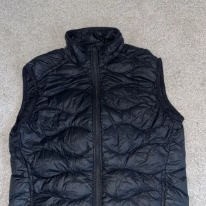 Svart quiltad väst från Peak Performance - Snygg svart quiltad väst från Peak Performance med dragkedja framtill. Perfekt för lager-på-lager under kyliga dagar. Västen har en stilren design med broderad logotyp på bröstet.