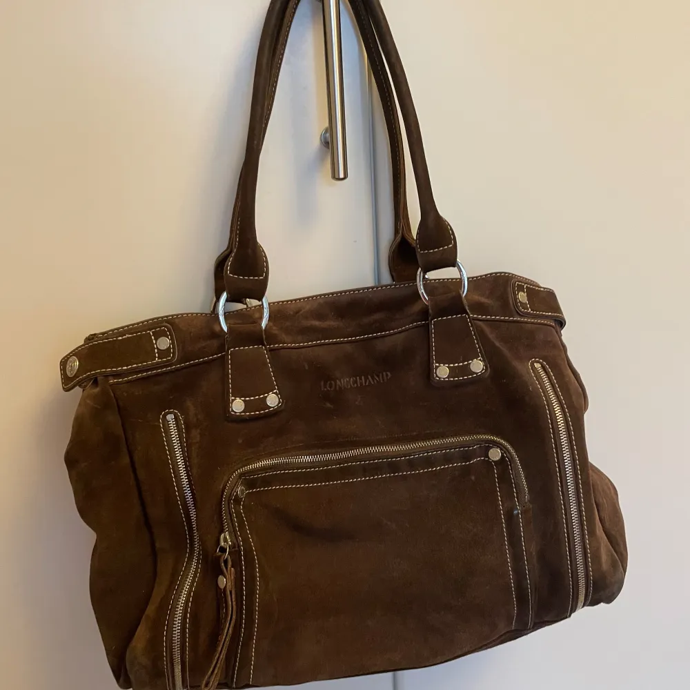 Snygg brun mockaväska från Longchamp med flera dragkedjor som kan användas för att göra väskan större och stiliga detaljer. Väskan har ett rymligt inre med ett lila fjärilsmönstrat foder. Den har en liten hake att fästa nycklar på, ett fack utanpå väskan och tre små fack inuti väskan. Perfekt för att bära med sig allt man behöver. Den har två handtag och en praktisk design. Självklart äkta. Man får plats med mappar från exacompta i den, så bra skolväska! Pris kan diskuteras!. Laukut & Käsilaukut.