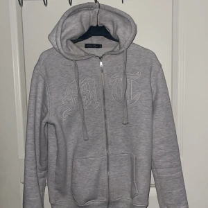 Tuttolente Zip Up Hoodie - Säljer en grå hoodie med dragkedja och tryck på både fram- och baksidan. Den har en justerbar huva med snören och en stor ficka framtill. Perfekt för en avslappnad stil.