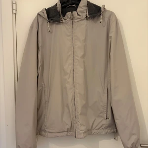 Beige vindjacka från Four Hands - Säljer en stilren beige vindjacka från Four Hands. Jackan har en dragkedja framtill och en praktisk huva. Perfekt för blåsiga dagar. Den är använd 1 gång 