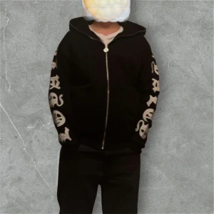 Svart hoodie med tryck - Säljer en svart hoodie med dragkedja och tryck i gotisk stil på ärmarna och huvan. Perfekt för en avslappnad stil med en cool edge. Den har långa ärmar och en bekväm passform.