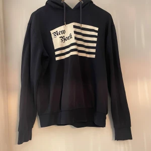 Mörkblå  hoodie från h&m - Säljer en svart mörkblå hoodie från Divided med tryck av texten 'New York' och ränder på framsidan. Det finns tecken på användning, trycket är lite slitet och ett hål finns på ärmen, men inget som inte går att fixa!