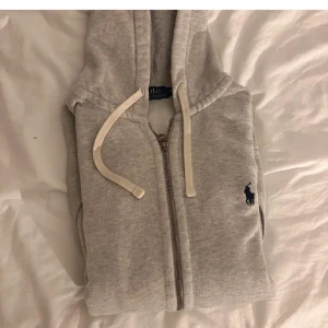 Grå hoodie från Ralph Lauren - Säljer en stilren grå hoodie från Ralph Lauren med dragkedja i bra skick har bara ett håll som man inte ser vid användning. Tveka inte att skriva om du har några frågor.