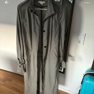 Rutig trenchcoat med bälte - Snygg rutig trenchcoat med knappar och bälte i midjan. Jackan har långa ärmar och ett klassiskt mönster i svart och beige. Perfekt för höst och vår.