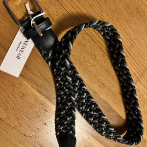 Svart och grått flätat bälte från New Wear - Snyggt flätat bälte från New Wear i svart och grått. Bältet har en klassisk metallspänne. Passar både till jeans och kostymbyxor. Säljer pågrund av felköp. Helt oanvänd och storlek 80