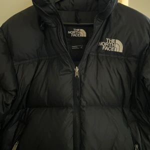 1996 RETRO NUPTSE JACKET UNISEX - The North Face - 1996 RETRO NUPTSE JACKET UNISEX - Dunjacka - black Säljer en svart dunjacka från The North Face med dragkedja och broderad logga på bröstet. Jackan har en hög krage och är perfekt för kalla dagar. Modellen är klassisk och tidlös, vilket gör den till ett måste i garderoben. 