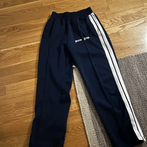 Blå träningsbyxor från Palm Angels - Snygga blå träningsbyxor från Palm Angels med vita ränder längs sidorna. Byxorna har en elastisk midja med snörning för bästa passform. Perfekta för en sportig look eller avslappnad stil. 