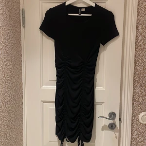 Svart klänning från H&M Divided - Snygg svart klänning från H&M Divided med rynkade detaljer och knytning framtill. Den har korta ärmar och en figurnära passform. Använd få tal gånger! 