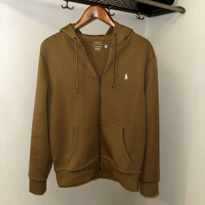 Beige hoodie från Polo Ralph Lauren - Säljer en stilren brun hoodie från Polo Ralph Lauren. Den har en dragkedja framtill och en broderad logotyp på bröstet. Perfekt för en avslappnad stil.