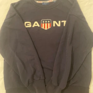 Mörkblå sweatshirt från GANT - Säljer en snygg mörkblå sweatshirt från GANT med broderad logga och sköld på bröstet. Tröjan har långa ärmar och ribbade muddar vid ärmslut och nederkant. Perfekt för en stilren och bekväm look.