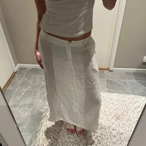 Vit långkjol - Säljer en stilren vit långkjol i ett lätt och luftigt material. Kjolen har en enkel design med dragkedja framtill men jag brukar använda den åt andra hållet. Den är både bekväm och elegant, vilket gör den till ett mångsidigt plagg i garderoben. Linnetyg och väldigt luftig. 