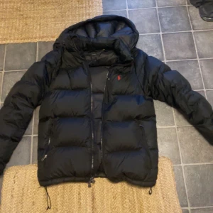 Svart pufferjacka från Ralph Lauren - Priset ej hugget i sten! Snygg svart pufferjacka från Ralph Lauren med dragkedja och broderad logga på bröstet. Jackan är perfekt för kyligare väder och har en klassisk design med långa ärmar. Finns en defekt som syns på bild 5 men annars helt perfekt skick🙌pris kan diskuteras!