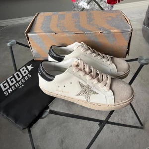 Helt nya Golden Goose Sneakers - Helt nya Golden Goose sneakers i vitt med svarta detaljer och en glittrig stjärna på sidan. Storlek 38. Box och dustbag medföljer. Boxen är lite skadad.