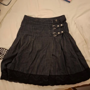 Vintage Jeanskjol från Lindex med spännen - Snygg jeanskjol från Lindex i mörkblå denim med dekorativa spännen och knappar framtill. Kjolen har en fin spetsdetalj längs nederkanten som ger en extra touch. Perfekt för en trendig look!