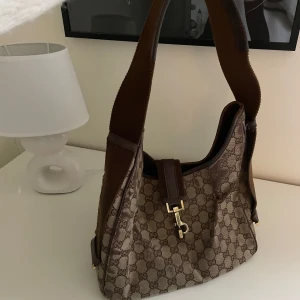 Brun mönstrad axelväska från Gucci - Säljer en snygg brun axelväska från Gucci med det klassiska monogrammönstret. Väskan har en axelrem och en elegant guldfärgad spänne framtill. Perfekt för att ge din outfit en lyxig touch.