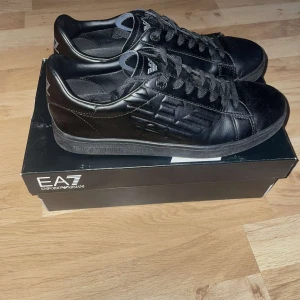 Svarta sneakers från EA7 - Snygga svarta sneakers från Emporio Armani med EA7-logga på hälen och tungan. Storleken är 42. Perfekta för en trendig look och passar till nästan alla klädstilar. Skorna är använda en gång under studenten sen har dem bara stått. Box medföljer också. Köpta på zalando. Hör av er vid frågor!!!! 😁