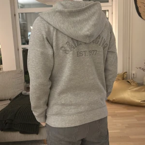 Grå Sail Racing zip  - Storlek M passar S, Säljer en grå hoodie från Sail Racing med dragkedja och tryck på bröstet och ryggen. Tröjan har en bekväm passform och är perfekt för kyligare dagar. Den har ribbade muddar vid ärmslut och nederkant.