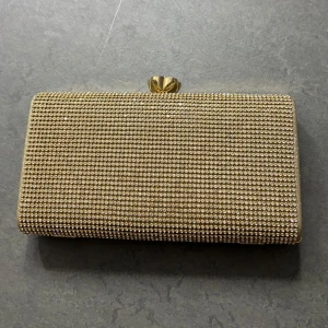 Glittrig guld kuvertväska - Snygg glittrig kuvertväska i guld med en elegant blomdetalj på låset. Väskan har en kedja för att bäras över axeln och en glittrig yta som fångar ljuset perfekt. Insidan är klädd i en ljusgul färg som ger en fin kontrast. Perfekt för att lägga till lite glamour till din outfit.