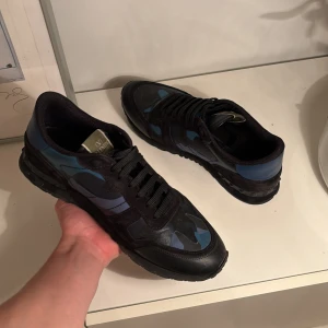 Valentino sneakers med blått kamouflagemönster - Snygga Valentino sneakers med ett unikt blått kamouflagemönster. Skorna har svarta snören och en robust sula för extra komfort. Perfekta för en stilren look. Använda ett par gånger men väldigt fint skick ändå.