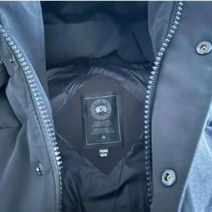 Svart dunjacka från Canada Goose - Säljer en svart dunjacka från Canada Goose med huva och dragkedja. Jackan har knappar och en logotyp på ärmen. Perfekt för kalla dagar. Aldrig använt 