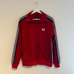Röd track jacket från Needles - Säljer en snygg röd track jacket från Needles med vita och mörka ränder längs ärmarna. Jackan har en dragkedja framtill och ett broderat fjärilsmärke på bröstet. Perfekt för en sportig look! Varan har endast används ett fåtal gånger.