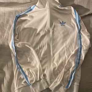 Vit träningsjacka från Adidas - Säljer en snygg vit träningsjacka från Adidas med ljusblå ränder längs ärmarna och det klassiska Adidas-loggan på bröstet. Jackan har dragkedja framtill och långa ärmar. Perfekt för en sportig look! Dm:a för mer bilder eller att diskutera pris 