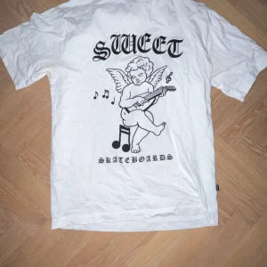 T-shirt - Säljer en vit t-shirt från Sweet Skateboards med ett coolt tryck av en ängel som spelar gitarr och musiknoter på framsidan. Perfekt för dig som gillar streetwear och skatekultur. T-shirten är kortärmad och gjord i bomull. Passar perfekt till vardags eller när du vill ha en avslappnad stil.