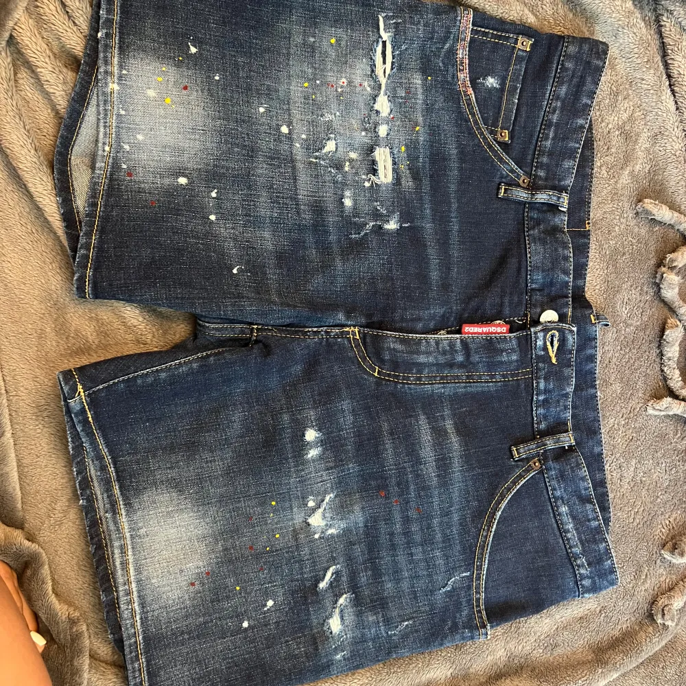 Säljer ett par snygga jeansshorts från DSQUARED2. De är mörkblå med coola slitningar och färgstänk som ger en unik look. Perfekta för sommaren och har en klassisk passform med fem fickor och knappgylf. Märket är känt för sin stil och kvalitet, så dessa shorts är ett måste för garderoben!. Shorts.