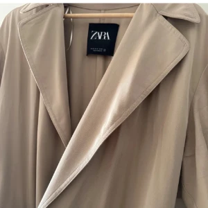 Beige trenchcoat från Zara - Säljer en superfin beige trenchcoat från Zara. Den har en klassisk design med ett bälte i midjan och långa ärmar. Perfekt för vår och höst när du vill se stilren ut. Jackan användt knappt två gg och passar till de flesta outfits!