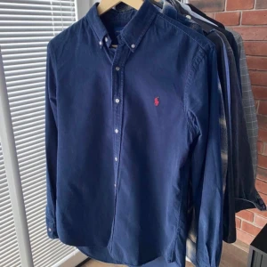 Mörkblå skjorta från Ralph Lauren - Säljer en snygg mörkblå skjorta från Ralph Lauren i storlek S. Skjortan är långärmad och har en klassisk krage med knappar hela vägen ner. Den är gjord av ett lätt och bekvämt material, perfekt för både vardag och fest. Den har också det ikoniska röda broderade logotypen på bröstet. Passar perfekt för en stilren look!