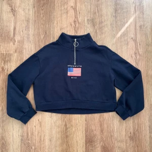 Mörkblå croppad tröja från H&M Divided - Säljer en snygg mörkblå croppad tröja från H&M Divided. Den har en cool dragkedja med ringdetalj vid halsen och ett tryck med texten 'Brooklyn NYC' och en amerikansk flagga på framsidan. Perfekt för en casual look!