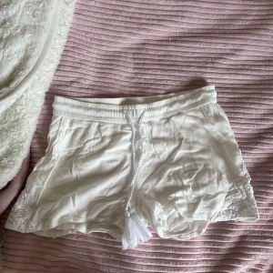 Vita shorts med spetsdetaljer - Säljer ett par supermysiga vita shorts med snygga spetsdetaljer på sidorna. De har en bekväm passform och är perfekta för sommardagar. Shortsen har en knytning framtill med tofsar som ger en bohemisk touch. Materialet känns mjukt och luftigt, perfekt för varma dagar! jätte fint mönster 