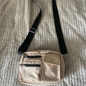 Beige axelväska med svarta detaljer - Säljer en beige axelväska med justerbar svart axelrem. Väskan har flera fack med dragkedjor på framsidan och ett stort huvudfack. Väskan är i syntetmaterial. Den har inga defekter. Pris kan diskuteras. Kontakta mig om du har fler frågor 😊