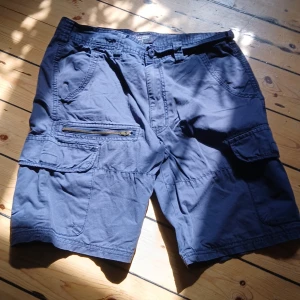 Blåa cargo shorts - Snygga blåa cargo shorts köpta i spanien för några år sedan, min brors gamla och har varit ganska välanvända men har bara legat i garderoben i några år så dam kan förekomma. Inga bilder på skickas. Midjemått rakt över 39cm, innrebenslängd 24cm💕