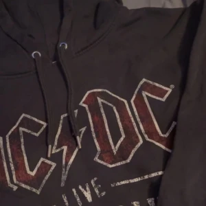 Svart AC/DC hoodie - Säljer en svart AC/DC hoodie från H&M. Den har ett coolt tryck med 'AC/DC Live World Tour' i rött och vitt på framsidan. Perfekt för rockälskare! Den är i storlek L och har en skön passform med långa ärmar och dragsko i huvan.