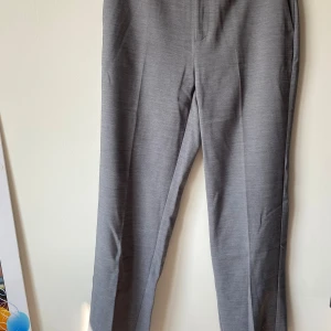 Grå kostymbyxor från Gina Tricot - Säljer ett par stilrena grå kostymbyxor från Gina Tricot i storlek 38. Byxorna har en klassisk rak passform och är perfekta för både kontoret och mer formella tillfällen. De är tillverkade i ett bekvämt material som ger en snygg och professionell look.
