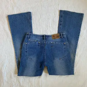 Säljer ett par snygga vintage blå bootcut jeans med  blommigt rhinestones på framsidan. Unika! Midjemått: ca 40 cm, innerbenslängd: ca 77 cm