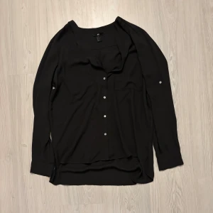 Svart blus från H&M - Snygg svart v-ringad blus från H&M i storlek M. Den har långa ärmar och knappar framtill, perfekt för både vardag och fest. Materialet känns lätt och luftigt, vilket gör den bekväm att bära hela dagen. Passar bra till både jeans och kjol! Obs Översta knappen saknas!