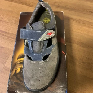 Grå skyddsskor från FTG - Säljer ett par grå skyddsskor från FTG i bra skick. Skorna har en robust design med kardborreband för enkel justering och har en slitstark sula för extra säkerhet. Perfekta för arbete där extra skydd behövs. Använda några gånger, finns bara målarfärgen på annars är i bra skick 