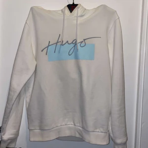 Vit hoodie från Hugo - Säljer en stilren vit hoodie från Hugo med ett coolt blått tryck på framsidan. Den har en klassisk passform med långa ärmar och en bekväm huva.  Hör av dig om du är intresserad!