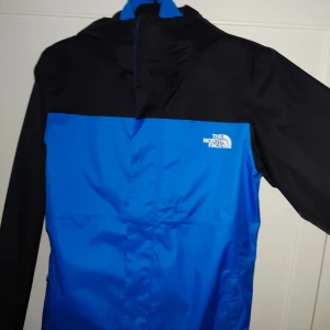 Blå och svart jacka från The North Face - Säljer en snygg blå och svart jacka från The North Face original med qr koden  2 in 1. Den har en dragkedja och är långärmad, perfekt för höst och vår. Jackan är i mycket bra skick och har en normal passform. Den är gjord av polyester och har en stilren design med The North Face-loggan på bröstet.