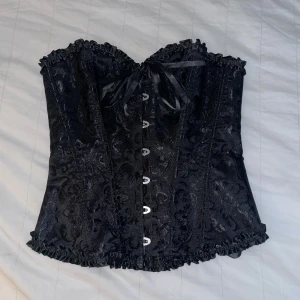 Svart korsett med spetsdetaljer - Snygg svart korsett med spetsdetaljer och knytning fram och bak. Den har en vacker glansig yta och volangkant upptill och nedtill. Perfekt för festliga tillfällen eller en utekväll. Passar bra till både kjol och byxor för en elegant look.